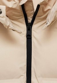 Giacca a vento beige con zip nera, bottoni a scatto sul colletto e tessuto liscio e lucido con una leggera lucentezza.