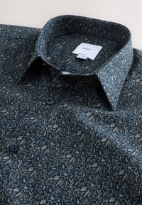 Tamnozelena košulja sa svijetlim cvjetnim uzorkom, ovratnikom s kopčama, dva gumba i tkanom etiketom. Materijal izgleda glatko s laganim sjajem.