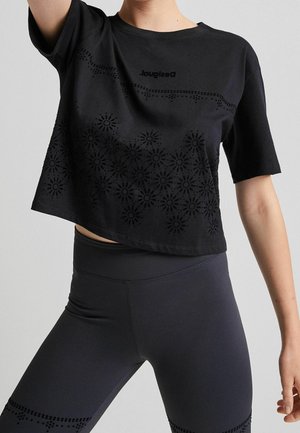 Frau trägt ein schwarzes Crop-Top mit Blumenmuster und schwarze hoch taillierte Leggings vor einem einfarbigen grauen Hintergrund.