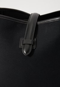 Bolso tote negro hecho de tela texturizada con un asa de cuero suave. Presenta un acento de cuero cosido cerca del punto de unión del asa.