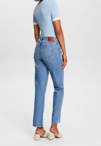 Jeans de tiro alto de color azul claro con piernas rectas, textura desgastada y parche de cuero marrón en la parte trasera de la cintura. Parte superior de manga corta blanca.