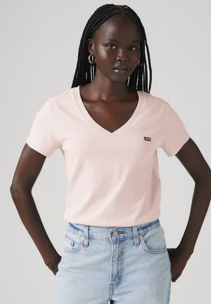 Mujer con el cabello trenzado que lleva pendientes de aro dorados, una camiseta de cuello en V rosa claro con pequeño logo y vaqueros de tiro alto azul claro, de pie con las manos en las caderas.