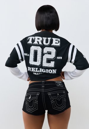 Osoba o krótkich czarnych włosach, nosząca czarno-biały long-sleeve crop top z "TRUE 02 RELIGION" oraz czarne szorty z białymi przeszyciami.