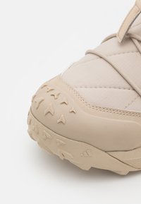 Zapatilla deportiva beige con una punta texturizada, parte superior de tela acolchada y suela de goma con un patrón que presenta pequeños tacos triangulares.