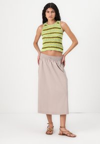 Jeune femme avec de longs cheveux bouclés portant un top en tricot à rayures vertes sans manches, une jupe midi beige à taille élastique et des sandales brunes à lanières.