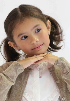 Kind maakt een beige cardigan met metallic vezels, een witte rimpelshirt en een zilveren hartketting. Haar in staarten, handen onder de kin.