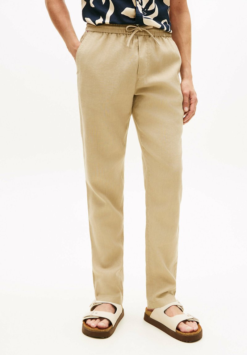 Personne portant un pantalon beige à cordon, une chemise à motifs noir et blanc, et des sandales beiges à deux brides avec des semelles marron.