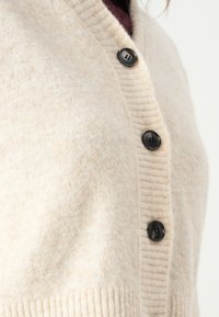 Beige cardigan laget av mykt, teksturert stoff, med en ribbestrikket krage og fire svarte knapper foran.