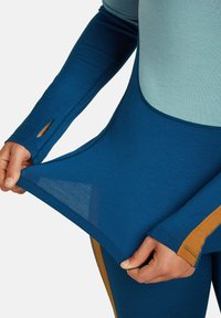 Una persona che tira l'orlo di una camicia a maniche lunghe blu e azzurro con strisce color senape sulle maniche e sui lati.