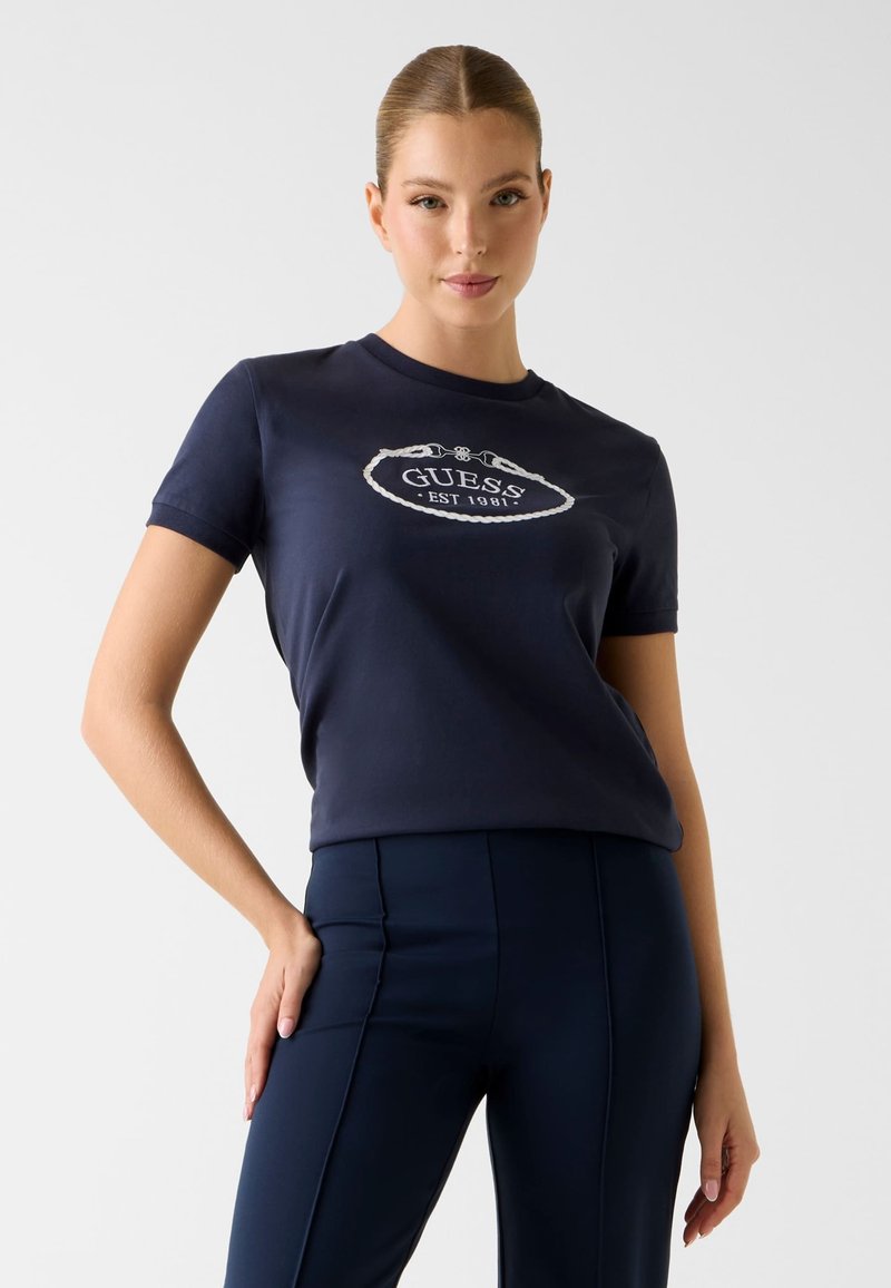 Granatowy bawełniany t-shirt z białym logo "GUESS" w formie liny. Krótkie rękawy i okrągły dekolt. W zestawie z ciemnymi spodniami.