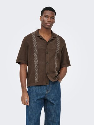 Mann mit kurzen lockigen Haaren, der ein braunes kurzärmeliges Hemd mit vertikalen Mustern und blaue Jeans trägt, steht mit einer Hand in der Tasche.