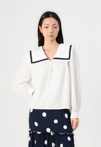 Hvit topp med stor krage med marineblå trim, slips ved halsen og lange pufferm. Båren over et marineblått prikkete skjørt.