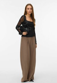 Kvinde stående med langt brunt hår, iført en sort gennemsigtig bluse med blomsterprint, brede brune bukser og med en sort clutch under armen.