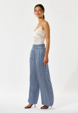 Femme aux cheveux bruns attachés en queue de cheval, portant un caraco en satin blanc, un pantalon large à motifs bleus et des sandales à talons noires, debout de profil.