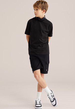 Barna hajú fiú áll, jobbra néz, fekete rövid ujjú pólót, fekete cargo rövidnadrágot, fehér boka zoknit és fekete-fehér tornacipőt visel.