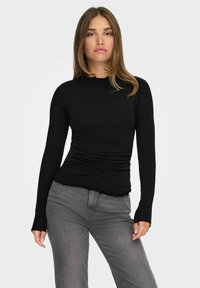 Top negro de manga larga con un diseño ajustado y textura arrugada en los laterales. Tiene un escote redondeado y puños acanalados, combinado con jeans grises.