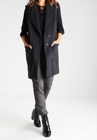 Gilet grigio scuro doppiopetto realizzato in tessuto spesso, con grandi tasche frontali, revers arrotondati e una vestibilità comoda, abbinato a pantaloni grigi.