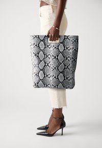 Black and white python print tote bag with a structured shape and an integrated handle. Smooth texture, paired with high-heeled black shoes.