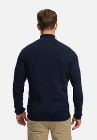 Marineblå kabelstrikket genser med høy krage, lange ermer og ribbestrikkede mansjetter, som viser et strukturert mønster over hele genseren.