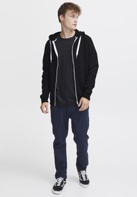 Sudadera negra con cremallera y cordones blancos, combinada con una camiseta negra y pantalones azules. Modelo lleva zapatillas negras. Fondo claro.