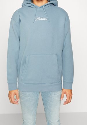 Persona que lleva una sudadera con capucha Hollister azul claro con bolsillo delantero y vaqueros de lavado claro, de pie frente a un fondo liso.