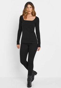 Mustat pitkät hihat -paita, jossa on neliön muotoinen pääntie, yhdistettynä mustiin istuviin housuihin ja nilkkakorkoihin. Kankaan pinta näyttää sileältä ja pehmeältä.