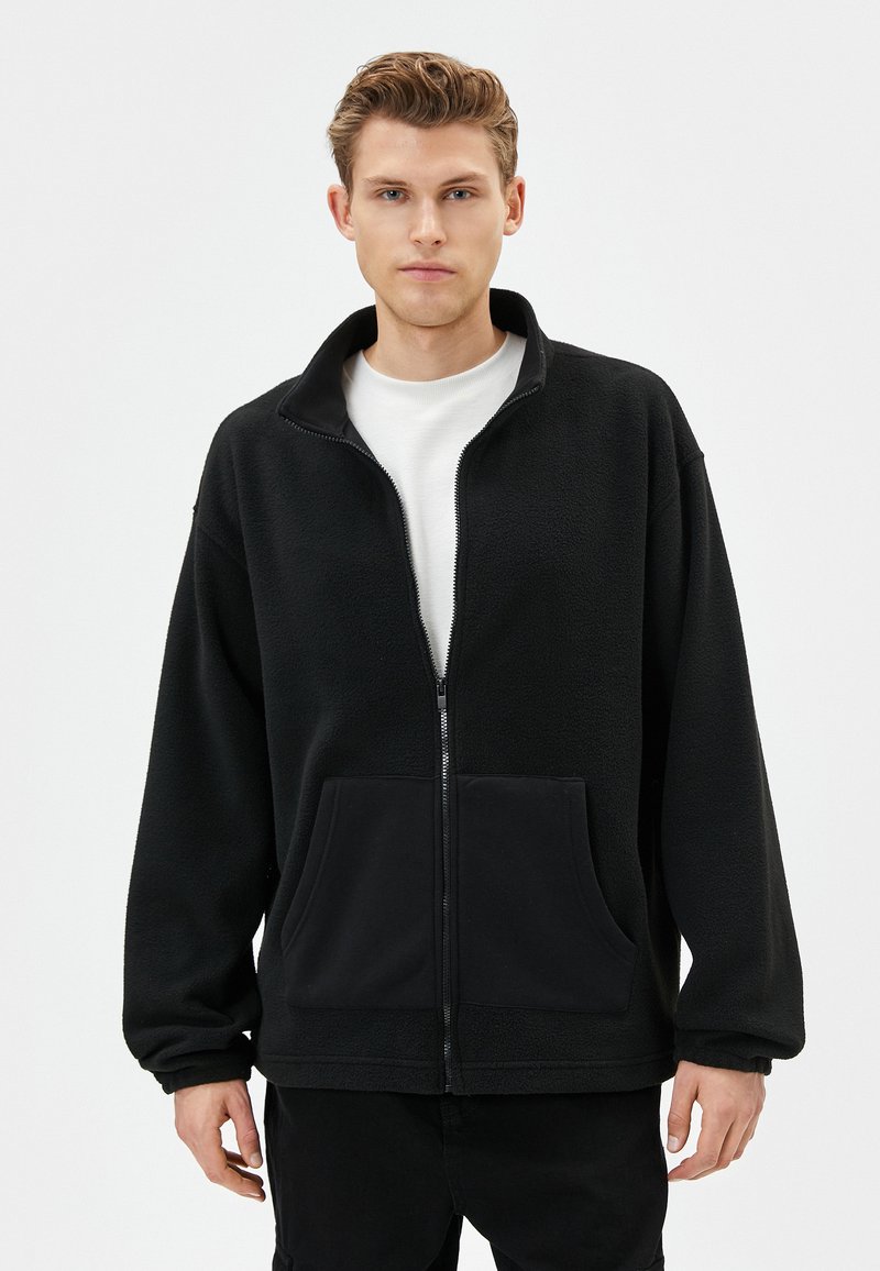 Chaqueta de felpa negra con cremallera frontal, cuello alto y dos bolsillos frontales. Presenta una textura suave y un corte relajado.