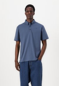 Polo majica s kratkimi rokavi v temno modri mešanici bombaža z ovratnikom, ki se zapenja na gumb in vezenim logotipom na levem prsnem delu. Modri jeans kratke hlače.