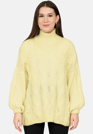 Junge Frau mit langen braunen Haaren, die goldene Creolen und einen blassgelben, grob gestrickten Pullover mit Ballonärmeln trägt.