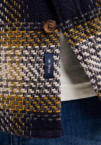 Cardigan lavorato a maglia in blu navy, giallo e bianco, con un bottone marrone, un motivo testurizzato e un'etichetta che recita "PETROL IND."