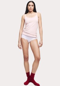 Lichtroze katoenen tanktop met kanten rand, gecombineerd met witte onderkleding en rode geribde sokken. Het model staat tegen een neutrale achtergrond.