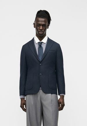 Uomo con dreadlocks vestito con blazer blu scuro, maglione grigio, camicia bianca, cravatta blu scuro e pantaloni grigi a pieghe, su sfondo semplice.
