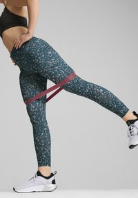 Legginsy w kolorze teal z teksturowanym, abstrakcyjnym wzorem; dopasowany krój; połączone z elastyczną taśmą oporową w kolorze bordowym. Białe buty sportowe.