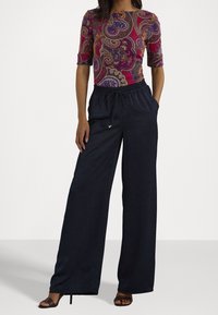 Mujer vestida con una blusa morada con un patrón paisley y mangas hasta el codo, pantalones anchos negros con cordón y sandalias de tacón negras.