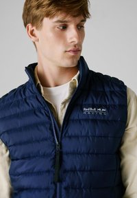 Marineblauwe puffervest met een hoge kraag, voorzien van horizontale gestikte lijnen en een rits aan de voorkant. Red Bull Racing-logo op de linkerborst.