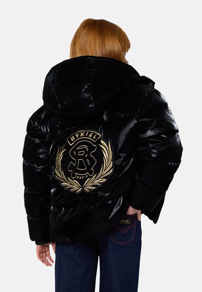 Personne aux cheveux roux portant une doudoune noire brillante avec un grand logo doré Rykiel Paris dans le dos et un jean bleu foncé.