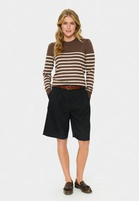 Donna che indossa un maglione a righe marrone e beige, pantaloni corti neri fino al ginocchio, una cintura marrone e mocassini di pelle marrone, in piedi con le mani nelle tasche.