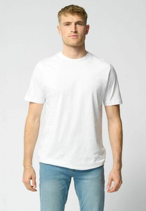 Lyshåret mand stående fremadrettet iført en ensfarvet hvid kortærmet t-shirt og lyseblå jeans mod en neutral baggrund.