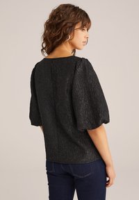 Čierna blúzka s textúrou, s nariasenými rukávmi, okrúhlym výstrihom a detailom v tvare kľúčovej dierky na chrbáte. Nosená s tmavými džínsami.