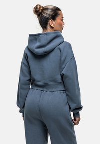 Felpa grigia a maniche lunghe con cappuccio ampio, spalle cadenti e polsini elasticizzati, abbinata a pantaloni della tuta coordinati con vita elastica.