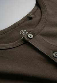 Gros plan sur l'encolure d'une chemise marron foncé avec des coutures, deux boutons et une étiquette de taille indiquant 6 ans.