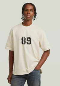 Crème-kleurige t-shirt met korte mouwen, voorzien van een grote zwarte opdruk "89" op de voorkant, gemaakt van zachte katoen.
