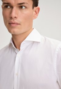 Witte overhemdd met een puntige kraag, voorzien van een gladde textuur en subtiele glans. Twee knopen zichtbaar bij de kraaglijn.