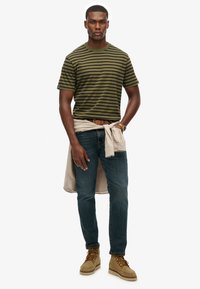 Olive green striped t-shirt, light beige shirt tied at the waist, dark jeans, tan boots, neutral background.