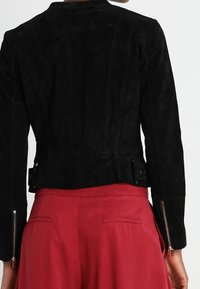 Svart mockajacka med en figursydd silhuett, med sömdetaljer på ryggen och dragkedjeärmar, i kombination med röda högmidjade byxor.