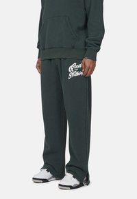 Donkergroene sweatpants met witte "Good for Nothing" tekst, van katoen gemaakt, rechte snit, relaxte pasvorm, gecombineerd met wit- en zwartkleurige sneakers.