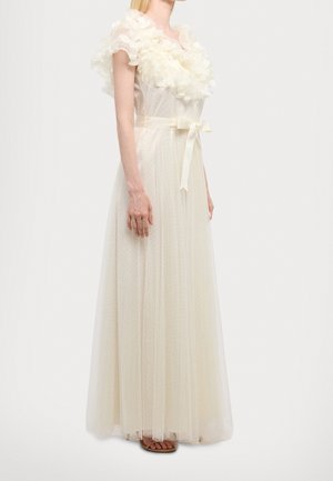 Frau trägt ein bodenlanges elfenbeinfarbenes Kleid mit floralen Rüschenärmeln, einem gepunkteten transparenten Überzug und einem Satinband, das in der Taille gebunden ist.