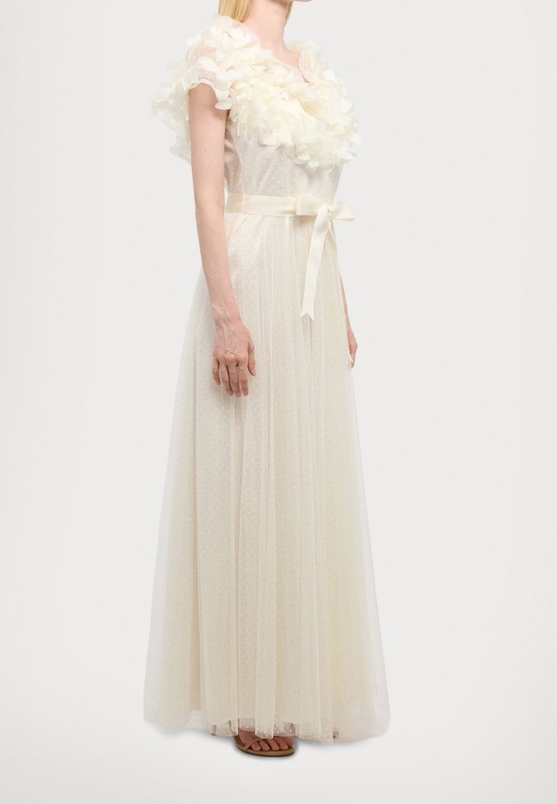 Femme portant une robe ivoire longue jusqu'au sol avec des manches à volants floraux, une surcouche transparente à pois et un ruban en satin noué à la taille.