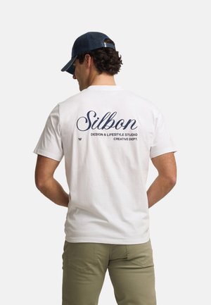 Hombre con una camiseta blanca con el texto "Silbon Design & Lifestyle Studio Creative Dept." en la espalda, una gorra azul marino y pantalones oliva.