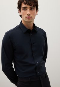 Camicia a maniche lunghe blu navy con colletto, caratterizzata da una texture liscia e chiusura con bottoni. Bottoni più chiari contrastano con il tessuto.